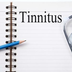 A notebook with lined paper and a spiral binding is open. The word 'Tinnitus' is written in large, bold letters at the top of the page. A blue pencil is placed diagonally across the notebook, and part of a stethoscope is visible on the right side of the image.
