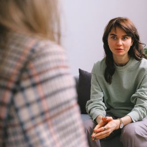 Two blurred individuals sitting indoors, facing each other during a conversation. One person wears a plaid shirt in the foreground, while the other in the background dons a light green sweatshirt. The setting is simple and neutral.