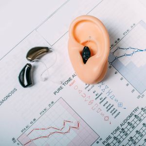 The image features a model of a human ear with a hearing aid inserted into the ear canal. It rests on top of a sheet of paper that displays audiometric graphs and data. Two additional hearing aids are placed next to the ear model. The audiometric graphs, labeled "Pure Tone Audiometry" and "Diagnosis," visually represent hearing test results.