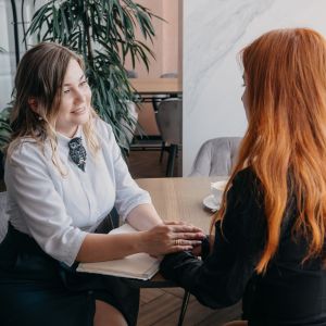 Two women sitting at a table having a supportive conversation, one holding the other's hands.